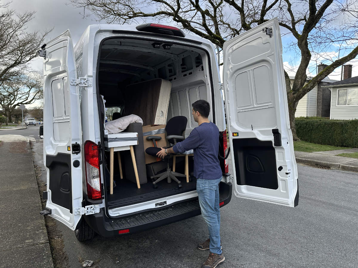 Man in a blue shirt securing a desk and office chair in the back of a white moving van on a Vancouver street.