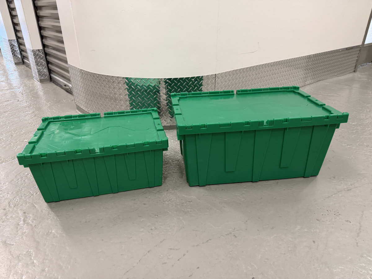 Two green plastic moving bins with lids on a concrete floor in a storage facility.