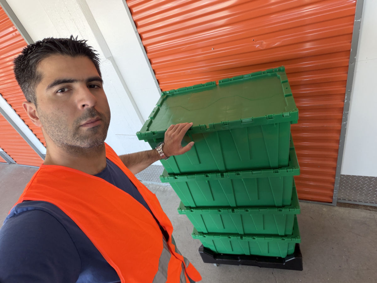 Mover in an orange safety vest taking a selfie next to four stacked green moving boxes on a dolly by orange storage doors.