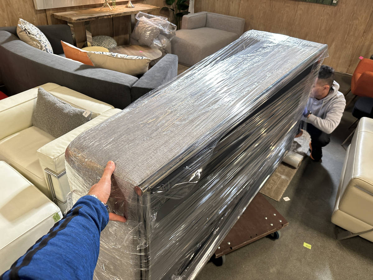 Wrapped sectional sofa being moved indoors by movers using a dolly, surrounded by living room furniture.