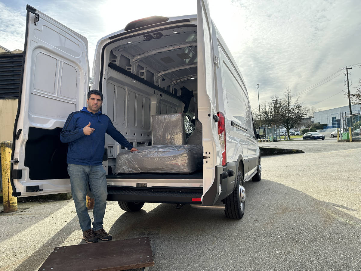 Man loading wrapped sofa into white cargo van with open doors, outdoor moving scene in daylight.
